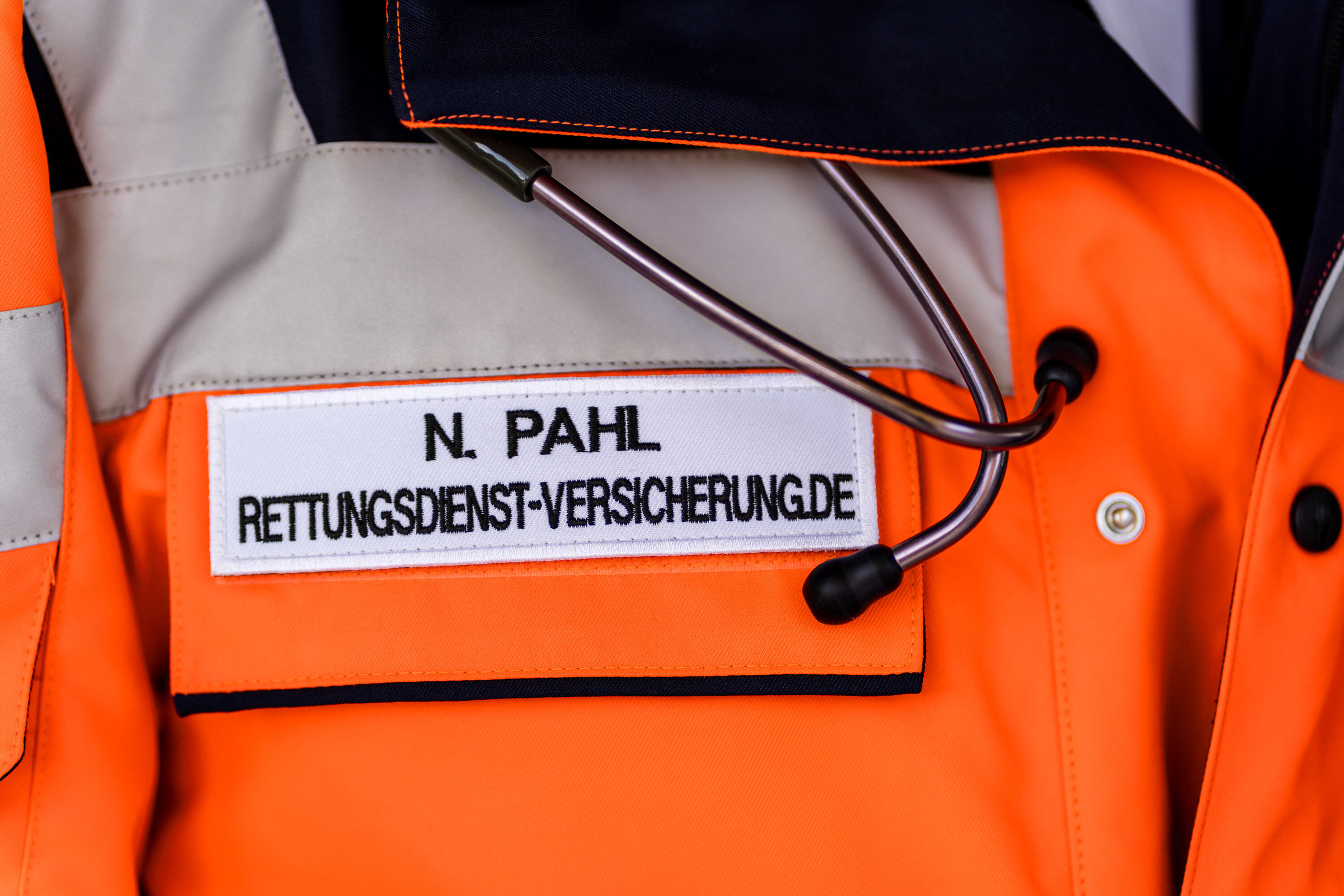 Namensschild N. Pahl, Rettungsdienst-Versicherung.de, auf einer orangen Einsatzjacke, daneben ein Stethoskop